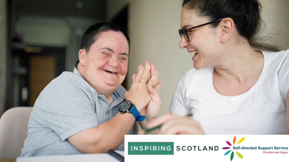 Woman with additional support needs sits with another woman. Both are laughing and smiling at each other while clasping hands.