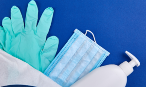 Blue background. In foreground is surgical gloves, PPE mask and handsanitiser bottle are laid out and fanned across the front.
